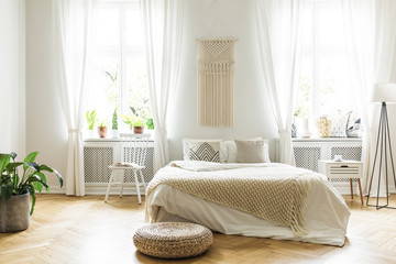 Pouf next to bed with blanket in white bedroom interior with plant and windows. Real photo - obrazy, fototapety, plakaty