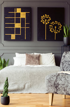 Bed with pillows under black and gold posters in bedroom interior with plants and chair. Real photo