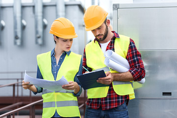 professional engineers in safety vests and hardhats working with blueprints and clipboard on roof