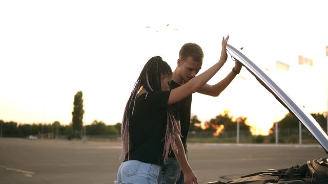 Side view of a young couple stopped while their road trip on the parking zone. Boy open up the car hood andpointing on the engine problem