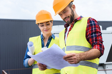 professional constructors in hardhats working with blueprints on roof