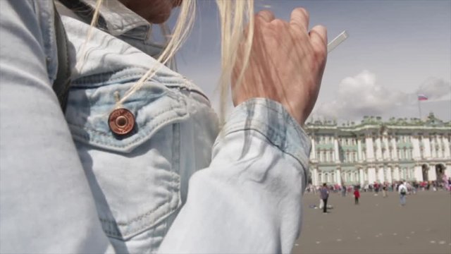 Young traveler blond woman wearing white dress, holding cellphone while standing near Hermitage on Palace Square in Saint-Petersburg and a large queue of people on the background standing on the