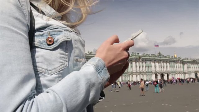Young traveler blond woman wearing white dress, holding cellphone while standing near Hermitage on Palace Square in Saint-Petersburg and a large queue of people on the background standing on the