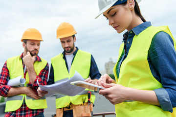 professional engineers in safety vests and helmets working with digital tablet and blueprints