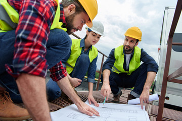 three engineers in hardhats working with blueprints on roof