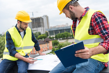 two engineers in helmets working with blueprints and clipboard on roof © LIGHTFIELD STUDIOS