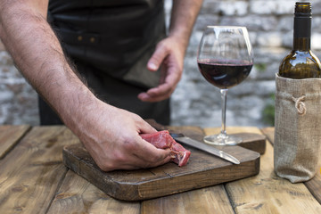 A glass of red wine on a wooden table. A man in a black leather apron prepares beef meat for dinner and a party. Copy space.