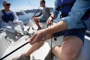 Abwaschbare Fototapete Segeln Nahaufnahme der männlichen Hände, die das Seil des Segelboots ziehen, während sie die Yacht festmachen und mit Freunden an Deck sitzen