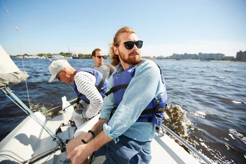 Photo sur Aluminium Naviguer Contenu jeune homme barbu insouciant en lunettes de soleil appréciant le yachting avec des amis : il est assis sur le pont et contemple le paysage marin
