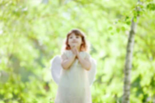 Blurred photo, background. Little Angel girl standing at the forest with arms close to her chest as in prayer.