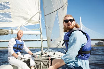 Papier Peint photo Naviguer Serious handsome men in life jacket frowning from sun looking around while traveling by sail boat on river