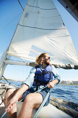 Serious pensive young bearded man with long hair wearing sunglasses holding ropes while sailing on...