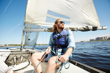 Fototapeta premium Serious pensive hipster young man with beard holding rope while operating sail boat and enjoying landscape around
