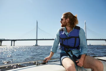 Photo sur Aluminium Naviguer Beau jeune homme sérieux et réfléchi avec barbe portant des lunettes de soleil et un gilet de sauvetage admirant le paysage aquatique assis sur le pont du bateau et tenant la balustrade