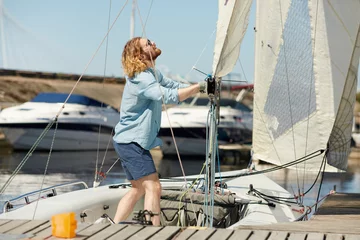 Photo sur Aluminium Naviguer Positive skilled young male sailor with beard wearing sunglasses adjusting sail on boat and examining it while fixing sail on boom