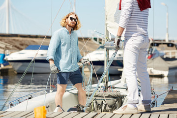 Serious handsome young man in sunglasses fixing rope on sail boat and consulting skilled sailor while preparing sailboat for tour