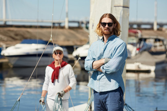 Serious hipster young bearded man in stylish sunglasses crossing arms on chest and looking at camera at yacht station, mature sailor with rope in background