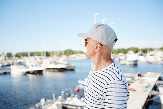 Pensive dreamy mature man in cap and sunglasses being on vacation looking at sailboats while standing on pier in yacht club