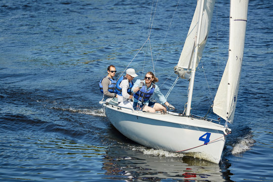 Group of free yachtsmen in sunglasses and life jackets sitting on sailboat deck and contemplating around while sailing at competition