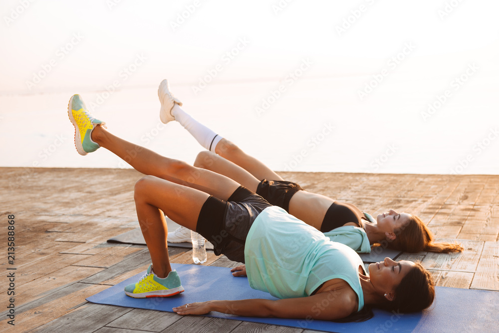 Wall mural young two sports women friends outdoors on the beach make sport exercises.