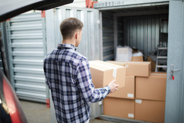 Rear view of young man in checkered shirt carrying packaged box from car to open container where...