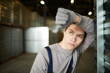 Restful pensive young guy working at distribution warehouse leaning on cargo container and looking...
