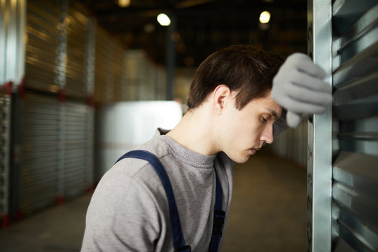 Exhausted young male cargo worker with closed eyes leaning on large container while tired from manual work in warehouse
