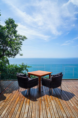 Table and rattan outdoor armchair setting on wooden deck and glass balcony with panoramic Andaman ocean sea view, green tree and blue sky background on sunny day