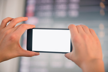 Mockup image of female hands holding black mobile phone with blank white screen over flight board in airport terminal