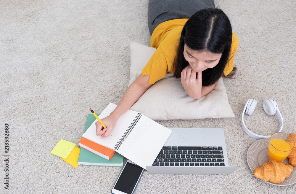 Poster top view asian woman lying on carpet and writing on notebook and learning on laptop with relax emoti