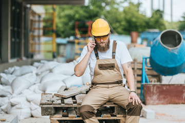 builder in hardhat and protective googles smoking cigarette and talking on smartphone at construction site