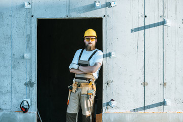  builder in protective googles and hardhat standing with crossed arms at construction site