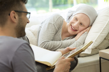Husband reading book to smiling sick woman with headscarf