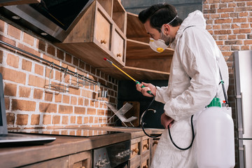 side view of pest control worker spraying pesticides under shelves in kitchen
