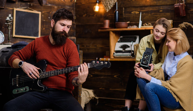 Bearded man with dreamy look playing guitar. Man with hipster beard meditating on music, tranquility concept. Musician enchanting women with lovely tunes. Girls recording performance on retro camera