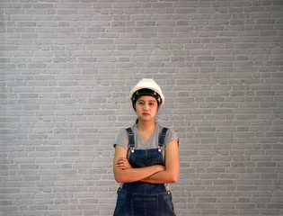 Technician woman ware white helmet with grey T-shirt and denim jeans apron dress standing straight angle and hugging chest on grey brick pattern background.