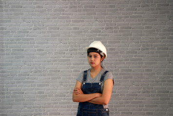 Technician woman ware white helmet with grey T-shirt and denim jeans apron dress standing forty five degree angle and hugging chest on grey brick pattern background.