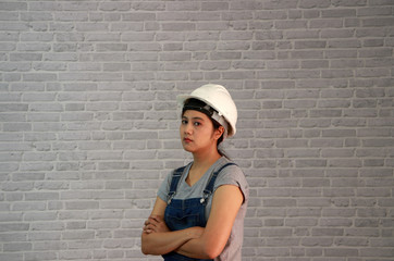 Technician woman ware white helmet with grey T-shirt and denim jeans apron dress standing forty five degree angle and hugging chest on grey brick pattern background.