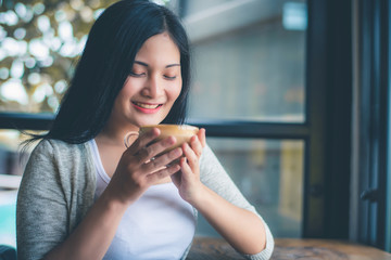 Beautiful attractive young Asian woman holding a cup of coffee in hand and sitting on sofa at cafe in the morning, vintage color tone. 