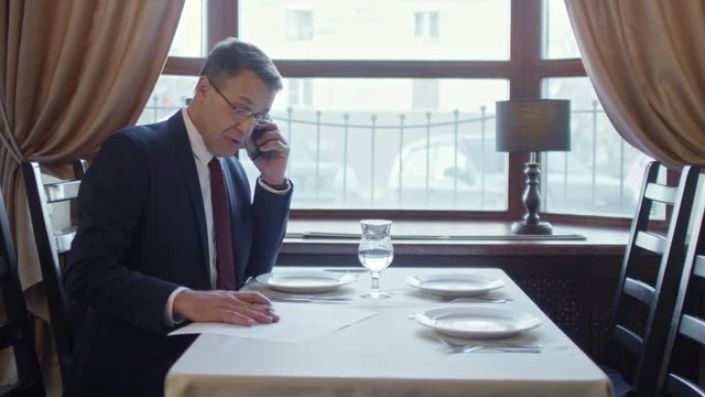 Zoom in shot of mature entrepreneur in business suit talking on the phone and writing in document when sitting at table by window at business lunch
