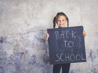 blond schoolgirl is holding blackboard with the words back to school in front of concrete background