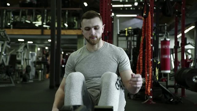 On rowing machine man in gym starts exercise on training. On sports equipment unshaven male sports person sits down and begins to perform work in the gym.