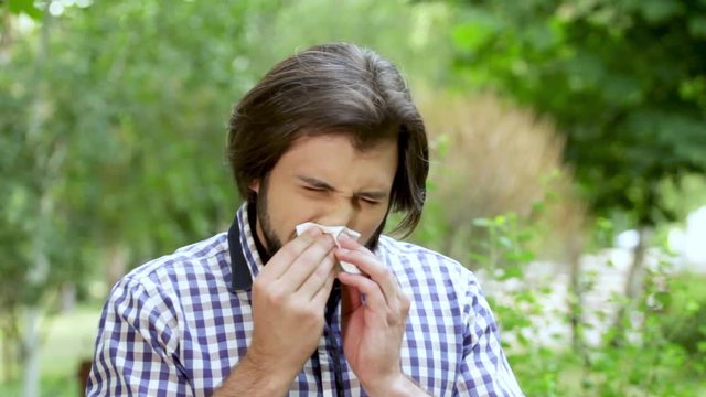 Man is standing in park. Guy is sneezing and covering nose with white napkin. After that he is mving with nose and sneezing again in napkin. Man is cleaning nose nad looking straight.