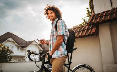 Young hipster male with curly hair looking to the camera, riding hthe city street, using his mobile...