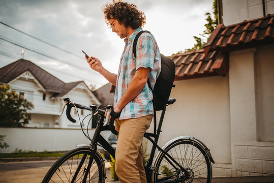 Side view of young happy male with curly hair looking on his mobile phone, choosing gps application for the road for bicycle. Happy smiling guy cyclist with backpack surfing internet using smart phone