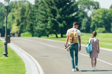 back view of teenage boy and girl with backpacks and skateboard walking together in park