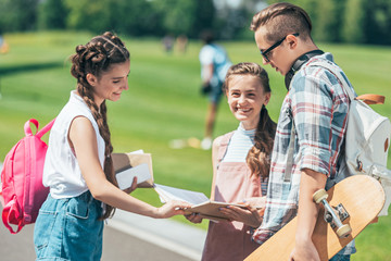 smiling teenage students with backpacks and skateboard standing and reading book together in park