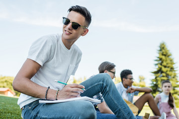 smiling teenage boy in sunglasses taking notes and looking away while studying with friends in park