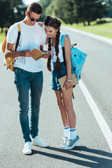 smiling teenage boy and girl with backpacks looking at notebook in park