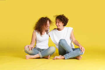Fototapeta na wymiar Two charming curly sisters or friends in white t-shirt and trendy jeans are sitting on studio yellow floor, hugging each other, spending time together fun. Family Relationships and Values
